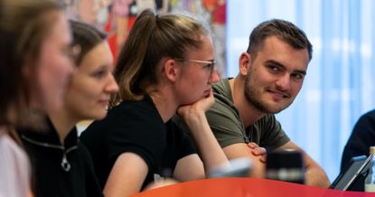 Group of young adults seated at a table in a discussion, with a woman resting her chin on her hand as she talks.