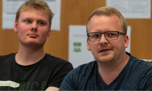 Two men seated indoors, one wearing glasses, looking toward the camera during a discussion in a classroom-like setting.