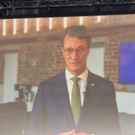 Man in a dark suit and green tie delivers a presentation on a stage, with a crest logo displayed on a screen behind him.