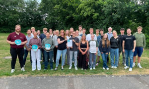 Group of about 25 people standing on a grassy park area, posing for a group photo; several hold blue circular signs.